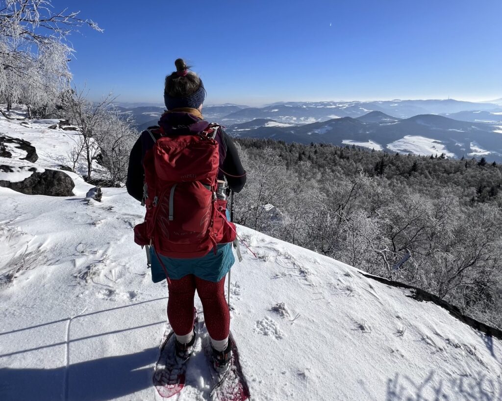 Teilnehmerin der Globetrotter Winterwandertage steht mit Schneeschuhen an der Kante des Gipfelplateaus des Hohen Schneeberges und beobachtet die Landschaft