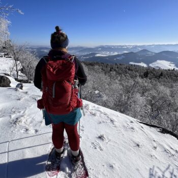 Teilnehmerin der Globetrotter Winterwandertage steht mit Schneeschuhen an der Kante des Gipfelplateaus des Hohen Schneeberges und beobachtet die Landschaft